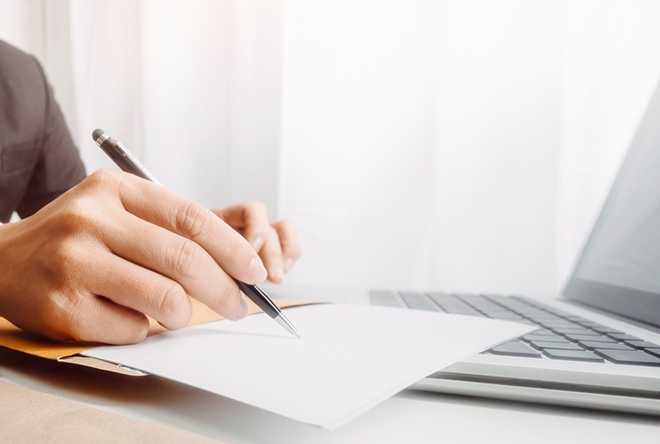 male with a pen writing on paper with a computer