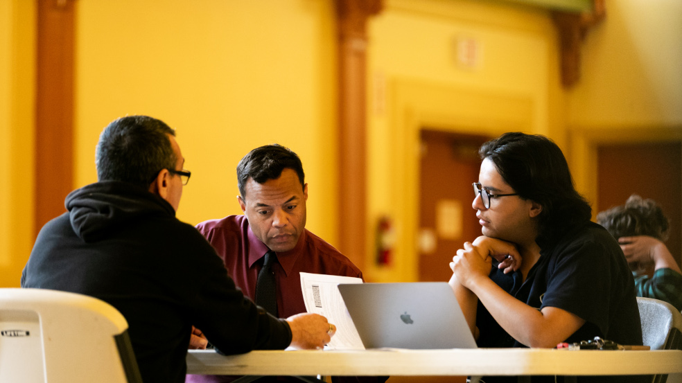 Three people sit at a table with a computer and paper forms.