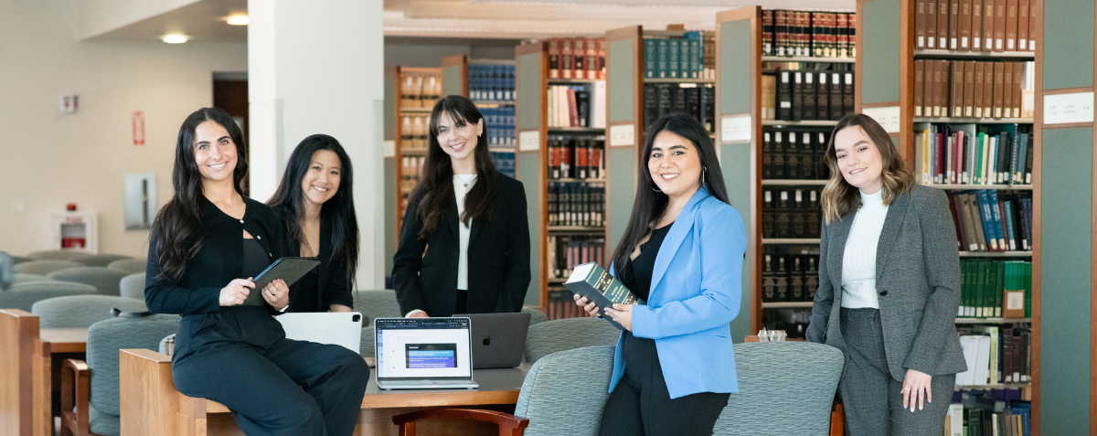 Five students stand around a table in front of bookshelves