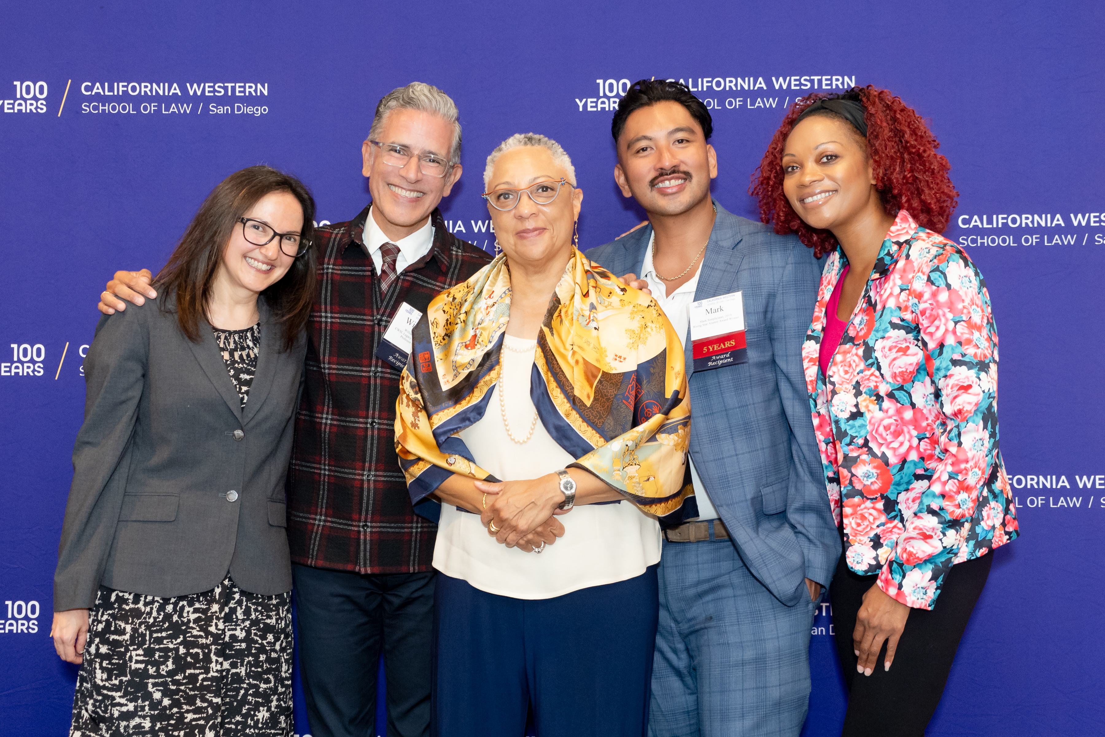 Dean Scott and the Distinguished Alumni Award Winners pose in front of a purple step and repeat. Left to right: Hon. Tila Nunn-Miller, Prof. William Aceves, Dean Sean Scott, Mark Simpliciano, Geneviéve Jones-Wright