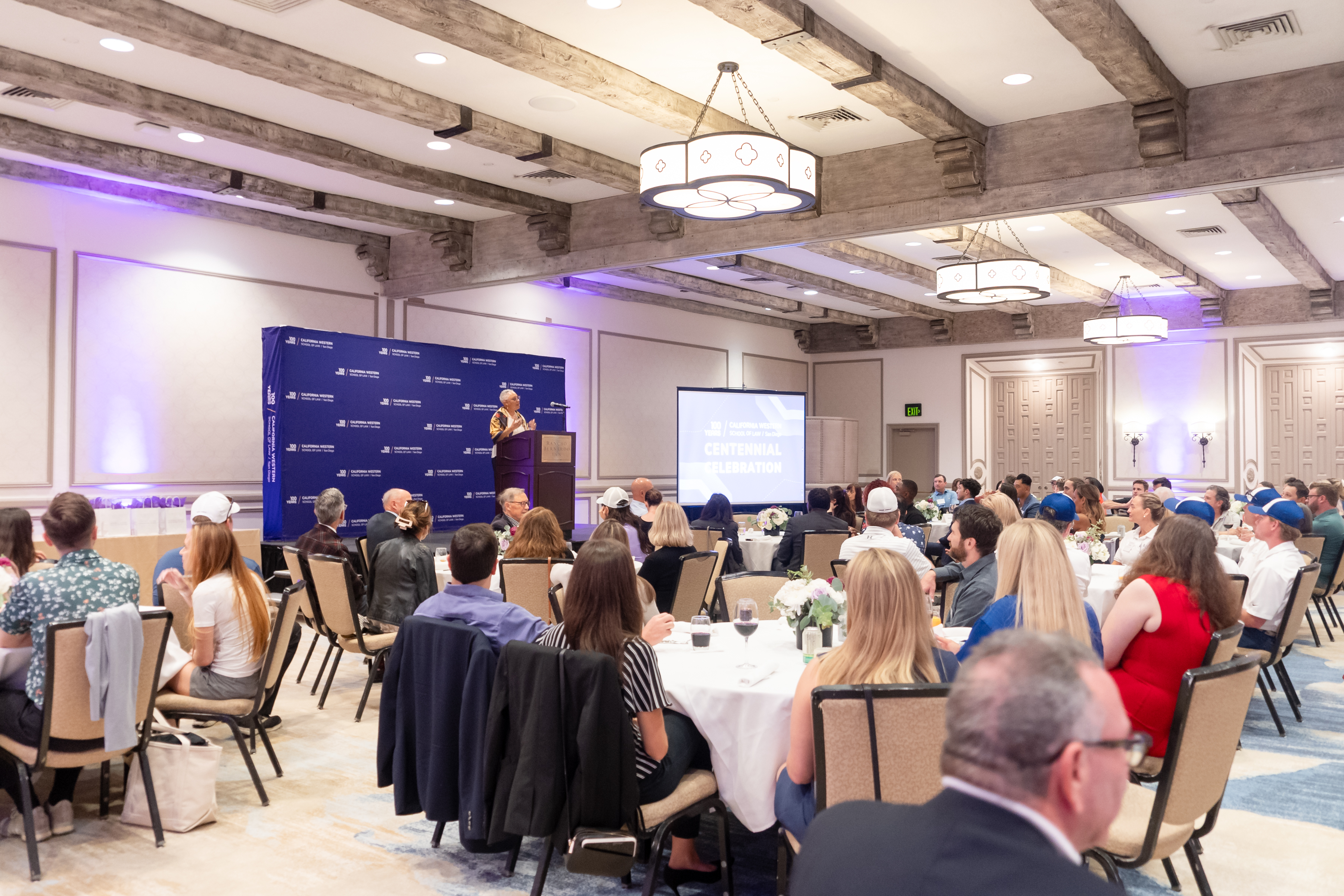 Dinner guests at round tables face the stage where Dean Sean Scott gives a speech from the podium with a purple step and repeat behind her. 