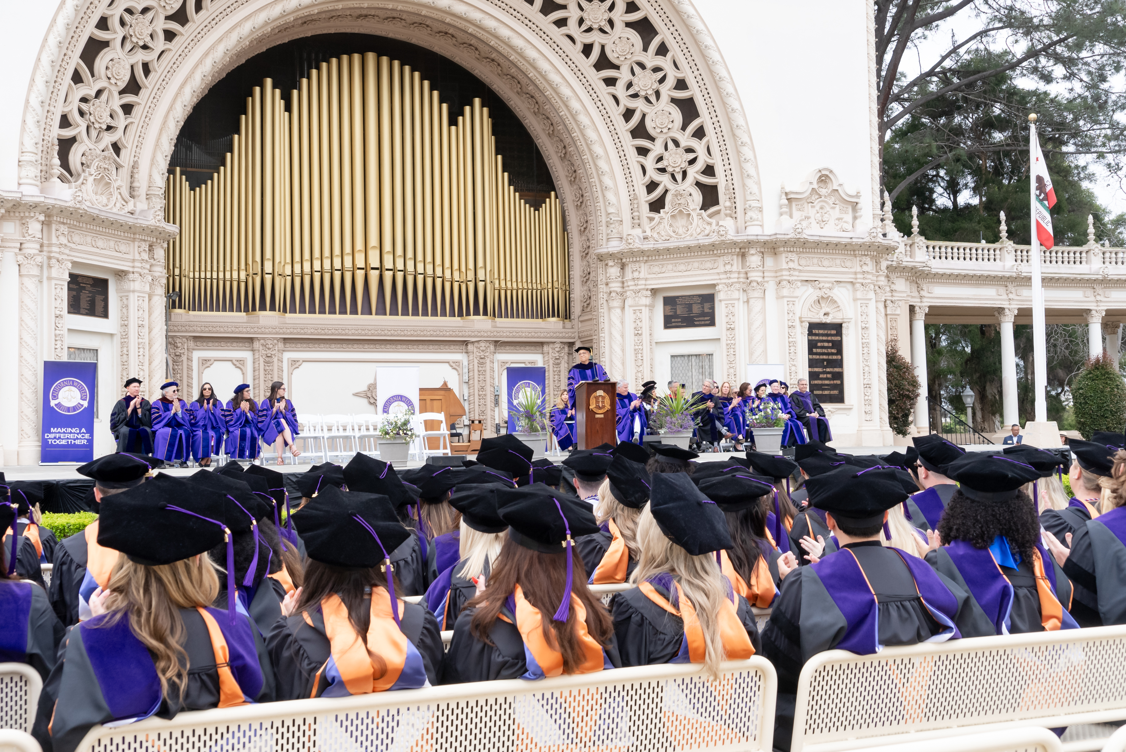 A crowd of graduates with purple and black JD regalia and orange, blue, and purple stoles face a stage featuring a pipe organ.