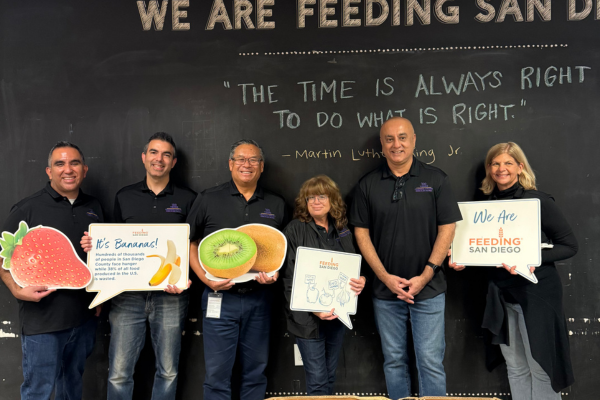 Six individuals in black polos and jeans stand with food cutouts and signs for Feeding San Diego.