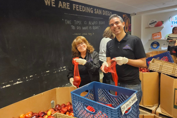 Two individuals in black polos wear gloves and prepare bags of fruit.