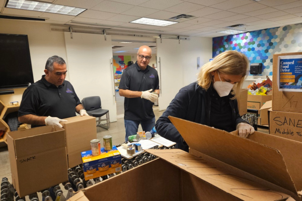 Three individuals in black polos unpack canned food from cardboard boxes.