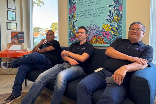 Three men in black polos and jeans sit on a blue couch.