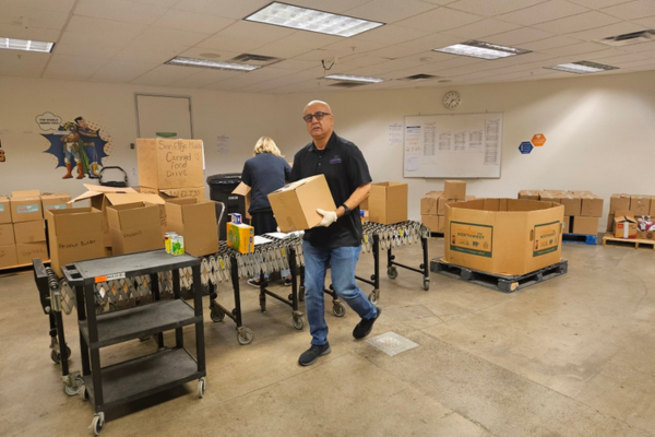 A man in a black polo and jeans carries a cardboard box in a room full of boxes.