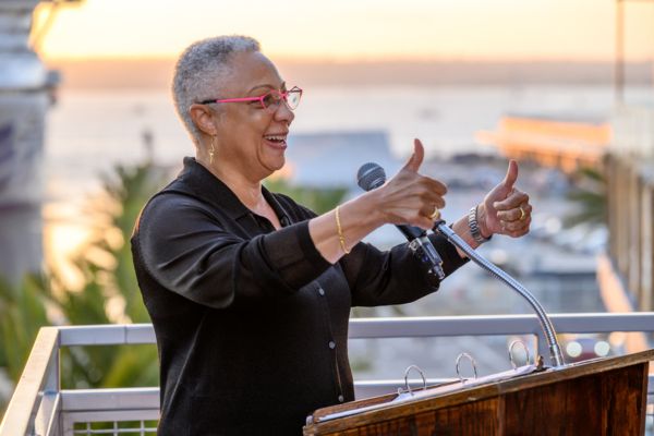 Dean Sean Scott wears pink glasses and a black blouse while standing at a podium with the sunset and harbor in the background. She smiles and holds two thumbs up.