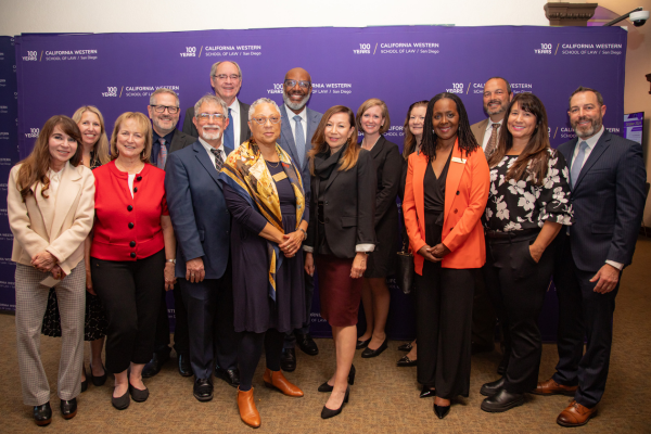 Sean Scott poses for a group photo with a large group of the judiciary in front of a purple step and repeat that features the centennial logo for CWSL.