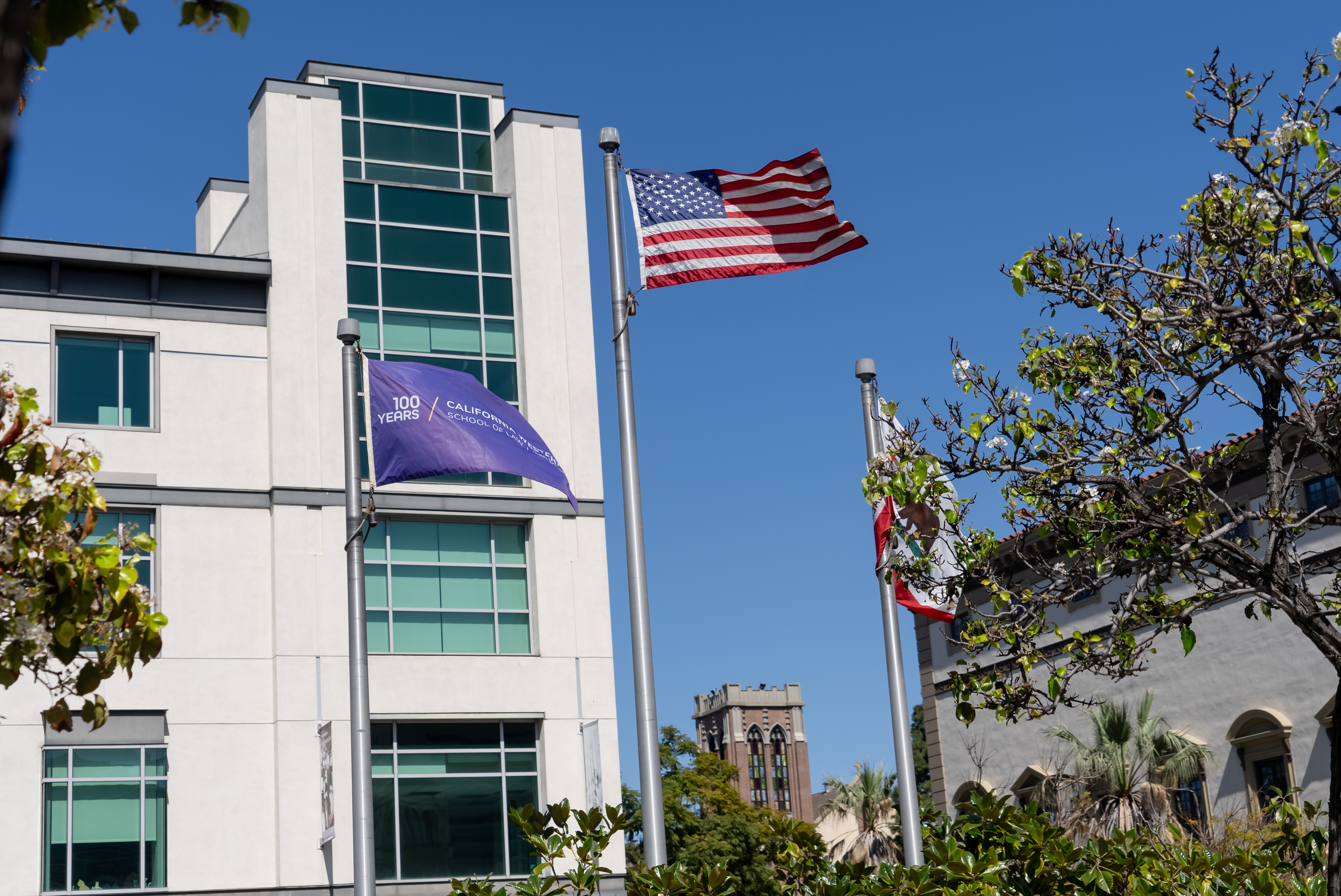 The American flag flies center and taller, with the purple centennial California Western flag to the left, and the California State flag, which is mostly blocked by a tree, to the right.