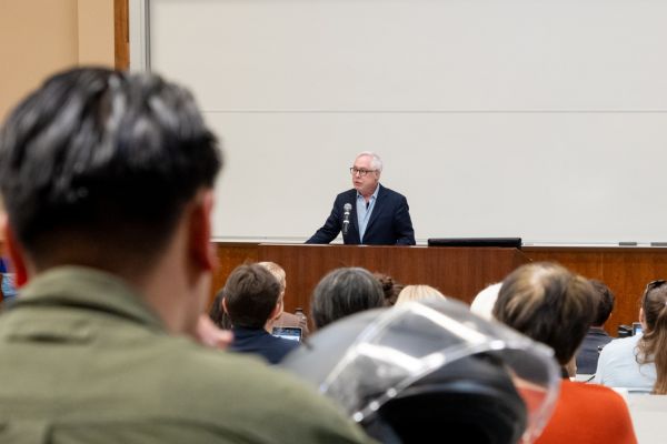 Louis Menand stands at the front of a full lecture hall wearing a navy suit and sky blue collared shirt.