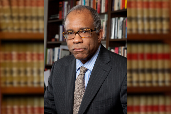 Against a background of books, Randall Kennedy wears a pinstripe suit and glasses for his headshot.