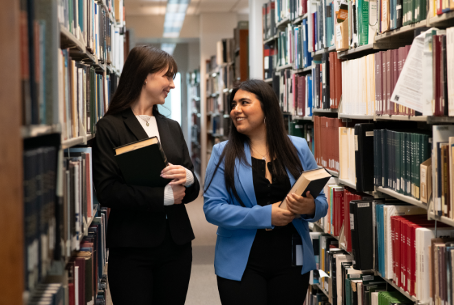 Two students carry their books through an aisle in a library