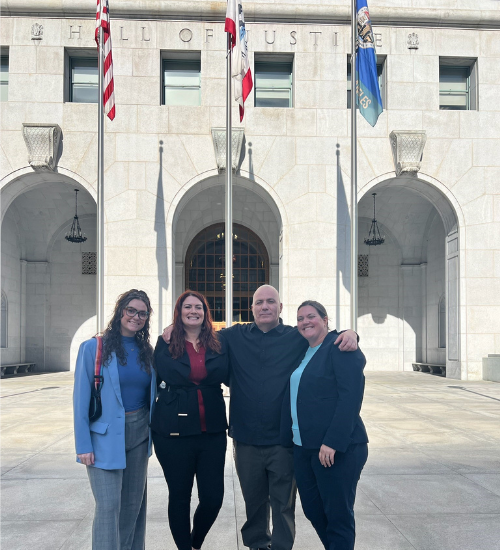 Four smiling individuals stand in foreground, in front of a courthouse entrance. The building's entrance has Hall of Justice engraved in large letters above the doorway behand three flag poles.