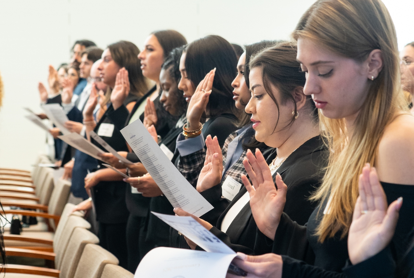 A group of professionally dressed individuals, mostly women, stand in a row with their right hands raised and holding papers in their left hands, reciting an oath or pledge. The setting appears formal, possibly a graduation, swearing-in ceremony, or professional induction event, with rows of chairs visible in the background.
