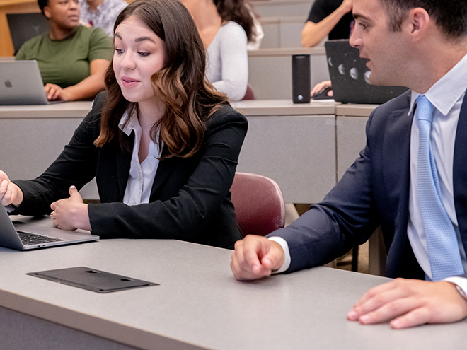 Two students in suits conversing while looking at a laptop in a lecture